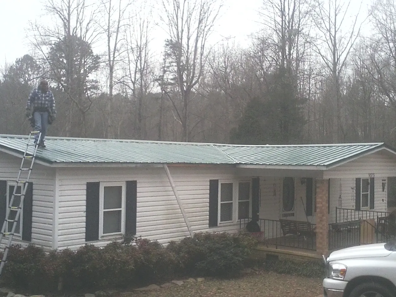 Worker on a ladder beside a new green metal roof on a ranch home in Enon