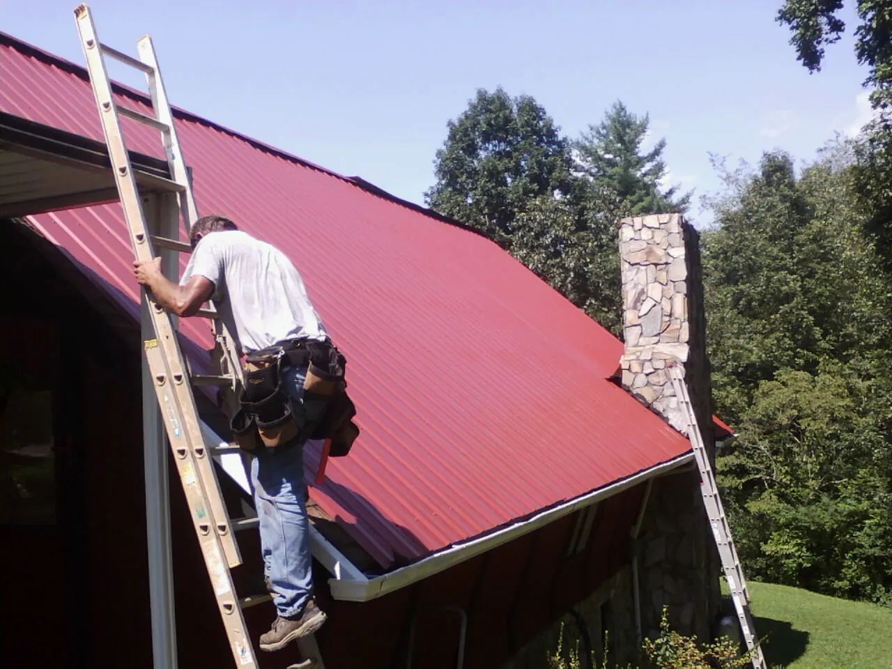 Roofer on a ladder beside a red metal roof and stone chimney — Soffit & Fascia Repair by Roofing Brothers in Enon